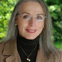 A woman with long gray hair and glasses, wearing a brown jacket over a black turtleneck and a pearl necklace, smiling with a blurred green background.