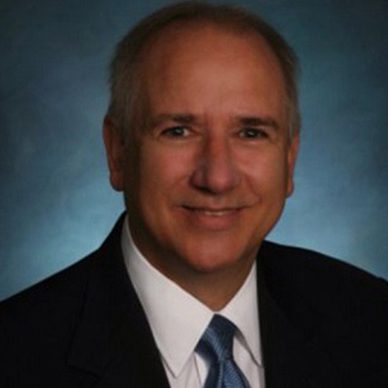 A middle-aged man with short gray hair, wearing a dark suit, white dress shirt, and blue tie, smiling at the camera in front of a dark, gradient background.